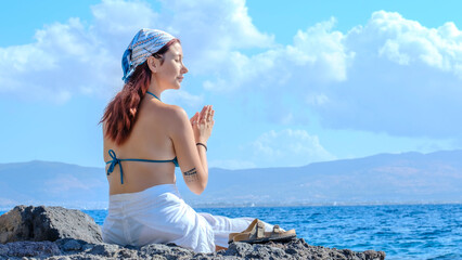 A young red-haired woman, wearing yoga pants and a bandana in her hair, is sitting on the rocks by...