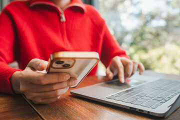 Close-up of a woman multitasking with a smartphone and laptop, wearing a red sweater, at a wooden table.