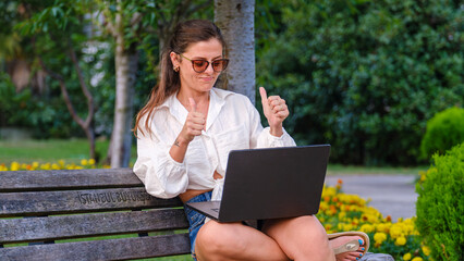 A beautiful middle-aged businesswoman sitting on a bench in a tree-lined park on a summer day,...
