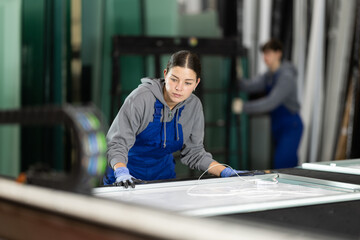 Fototapeta premium Girl glazer employee in blue overall works in glass processing shop. She carries out installation work, products for renovation of residential premises made of glass and plastic..