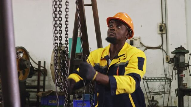 African American factory worker wearing hard hat and gloves pulls chain on steel beam for safety, while lifting and moving heavy machinery placed on floor in automotive parts lathe workshop.