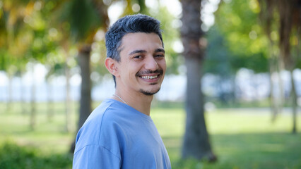A dark-haired young man smiling at the camera in a tree-filled park