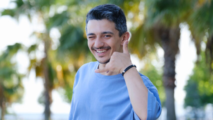 A dark-haired young man in a tree-filled park on a summer day, looking at the camera, making a...