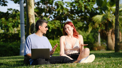 A young business woman and man working happily on their laptops while leaning on a tree trunk in a...