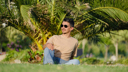 A handsome, young, dark-haired man lounging on the grass in a tree-filled park on a summer day and striking cool poses while enjoying himself