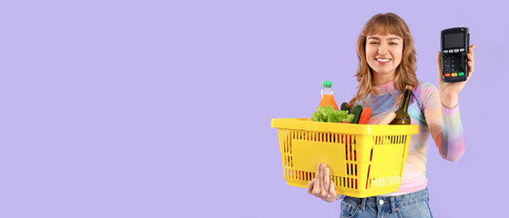 Young woman with full shopping basket and payment terminal on lilac background