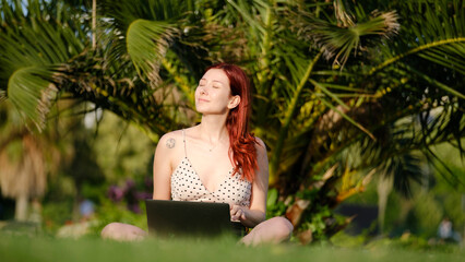 A beautiful, young, red-haired businesswoman sitting on the grass in a tree-filled park, happily...