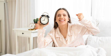 Happy young woman with alarm clock in bedroom