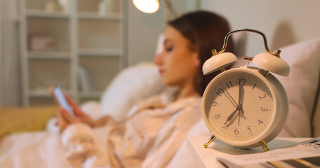 Alarm clock on table of young woman using mobile phone in bedroom at night, closeup