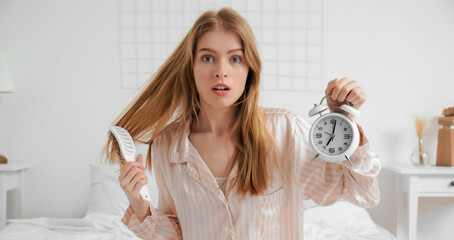 Shocked young woman holding alarm clock and hairbrush in bed at home