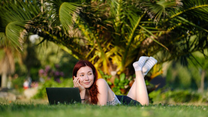 A beautiful, young, red-haired businesswoman sitting on the grass in a tree-filled park, happily...