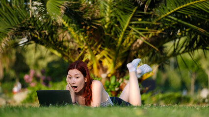 A beautiful, young, red-haired businesswoman sitting on the grass in a tree-filled park, happily...