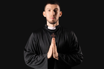 Young priest with cross praying on black background