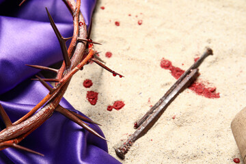 Crown of thorns with purple fabric, nails and blood on sand background, closeup