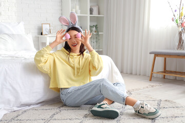 Young Asian woman with bunny ears and Easter eggs sitting on floor in bedroom