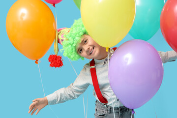 Funny little boy in clown wig and hat with balloons on blue background. April Fools Day celebration