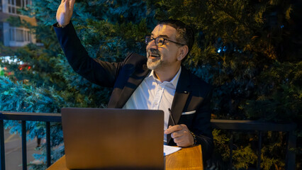 Mature handsome businessman in suit cheerful greeting someone he sees working on laptop outside...