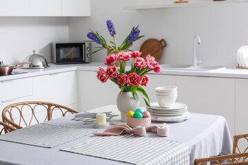 Vase with tulips, Easter eggs and dinnerware on dining table in kitchen