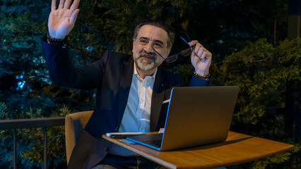 Mature handsome businessman in suit cheerful greeting someone he sees working on laptop outside...