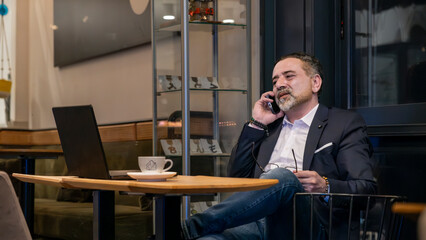 Handsome mature businessman working with laptop outside office environment, making phone call