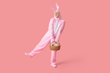 Happy young man in Easter bunny costume holding basket on pink background