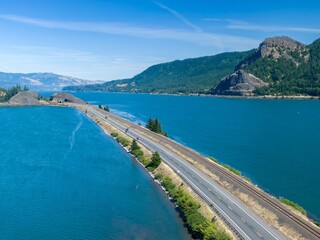 Naklejka premium Highway and train tracks alongside a scenic Columbia River. Cars travel along the road, showcasing the beauty of the Columbia River Gorge. ,Drano, Washington, USA