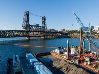 Riverfront dredging operation near a historic Steel Bridge. Barges and cranes remove sediment....