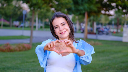 A young, beautiful businesswoman with brown hair is looking at the camera while making a heart...