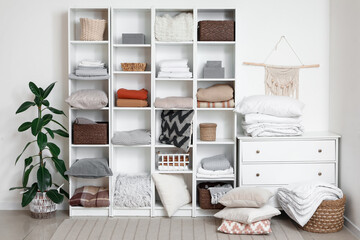 Shelving unit with clean bed sheets, plaids and baskets near white wall in interior of room