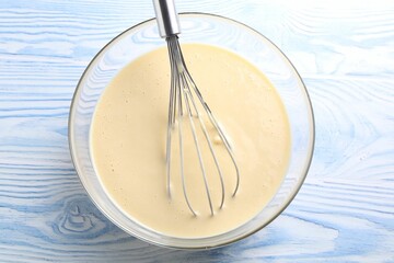 Whisk and bowl of dough on light blue wooden table, top view