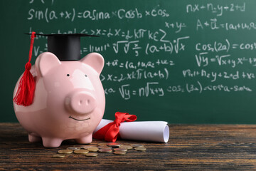 Piggy bank with graduation hat, coins and diploma on table near green blackboard. Student loan...
