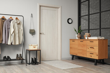 Interior of modern hall with beige door, chest of drawers and rack with clothes near grey wall