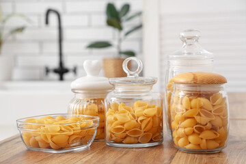 Jars and bowl with raw pasta shells on table in kitchen