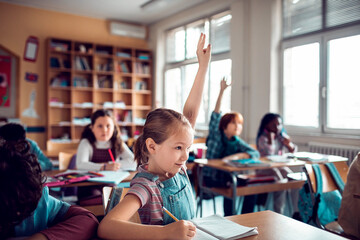 Young Caucasian girl raising her hand for the teacher while at the elementary school classroom