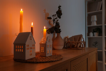 House shaped holders with burning candles on chest of drawers in dark room