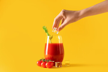 Woman putting ice cube into glass of bloody mary with celery, tomatoes and ice cubes on yellow...