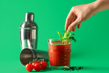 Woman putting celery into glass of bloody mary with tomatoes and peppercorns on green background