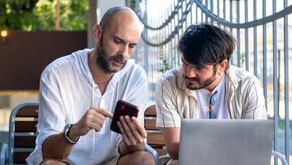 Two middle-aged male professionals are happily working on their laptops in the garden of a café on...