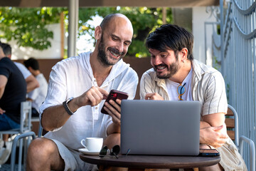Fototapeta premium Two middle-aged men professionals happily work on their laptops in the garden of a cafe on a summer day