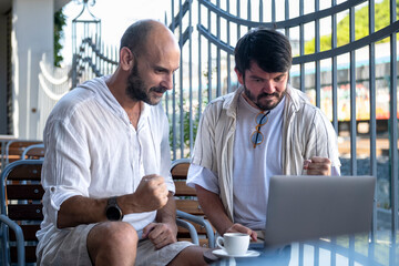 Two middle-aged male professionals are happily working on their laptops in the garden of a café on...