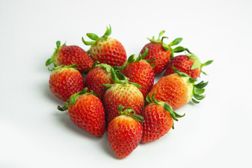 strawberries are arranged in a heart shape on a white background. The berries are red with green stems, some showing a transition to lighter, unripe areas. The image is a top-down view