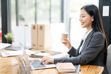 Happy young Asian businesswoman using laptop while analyzing reports in office.