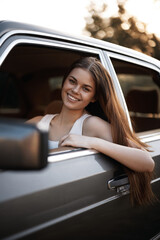 Happy young woman with long hair smiling from a car window in a sunny outdoor setting, wearing a casual white tank top and enjoying a relaxed moment.