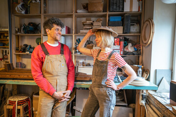 Charming carpenter couple tries on handmade wooden lampshade as hat for joke in workshop. Family business, funny break at work and love for manual work, woodworking creativity, craftsmanship bond