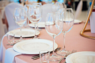 Table setting with light pink tablecloth close up. Wine and beverage glasses, white plates, forks and knives, preparation of the restaurant for the reception of guests