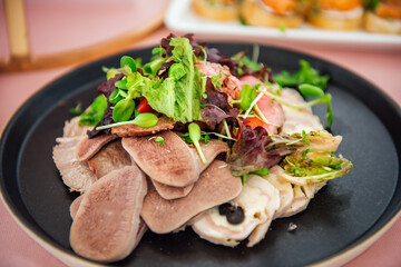 A variety of meat snacks, boiled beef tongue, roast beef and meat roll on a black plate with lettuce leaves and microgreens. Restaurant dish on a light pink tablecloth
