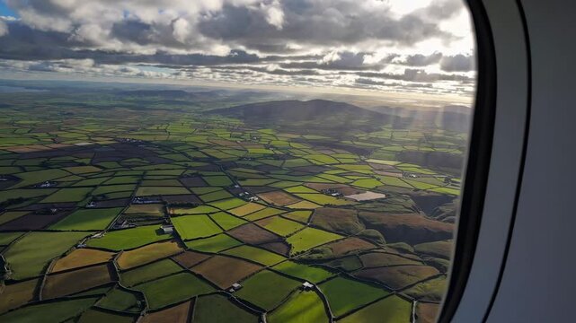 Aerial view from an airplane window showing a patchwork of green fields under a cloudy sky, capturing a serene landscape video from a high angle.