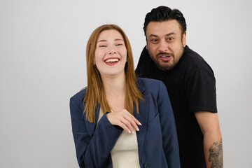 A young businesswoman in a suit and a middle-aged man wearing a black t-shirt stand in front of a white background in a studio environment, making funny facial expressions and looking at the camera