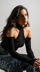 In a studio setting with a white background, a young black-haired woman wearing a black blouse and jeans strikes an artistic pose