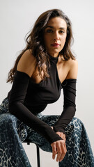In a studio setting with a white background, a young black-haired woman wearing a black blouse and jeans strikes an artistic pose